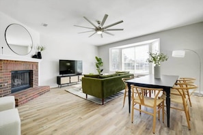 Een moderne woonkamer met een open haard, een groene bank, een eettafel met stoelen, een plafondventilator en een groot raam.