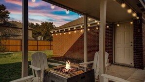 A covered patio with a fire pit, two Adirondack chairs, and string lights. - Lewisville Estate - Spacious 5BR w/ Game Room (Lewisville)