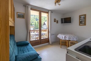 A room with a sofa, a table with a floral tablecloth, a TV mounted on the wall, and a sliding glass door leading to an outdoor area with a deck and a building.