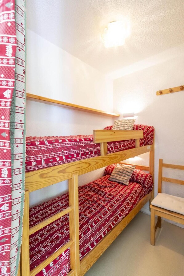 A bunk bed with red patterned bedding, a wooden chair, and a wall-mounted light fixture.
