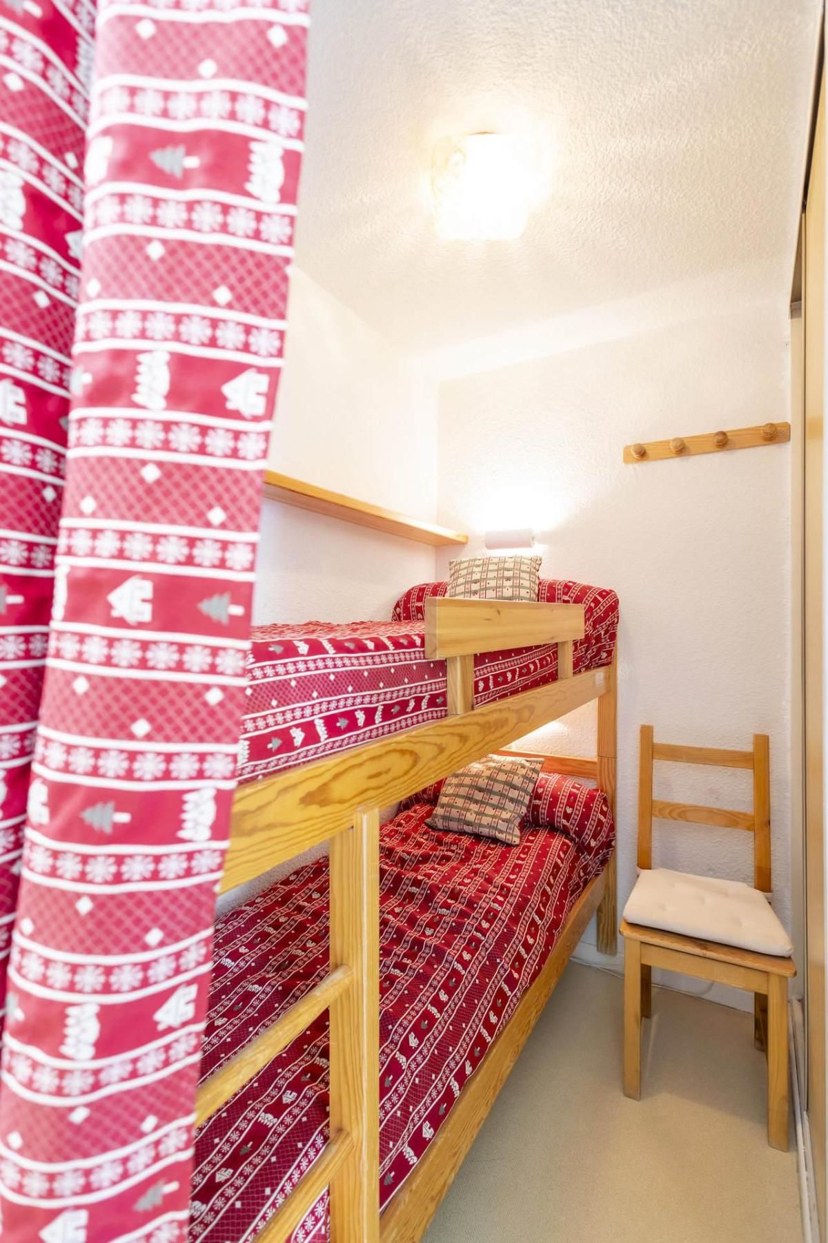 En køjeseng med et rødt mønstret sengetæppe, en træstol og et gardin med et rødt og hvidt design.