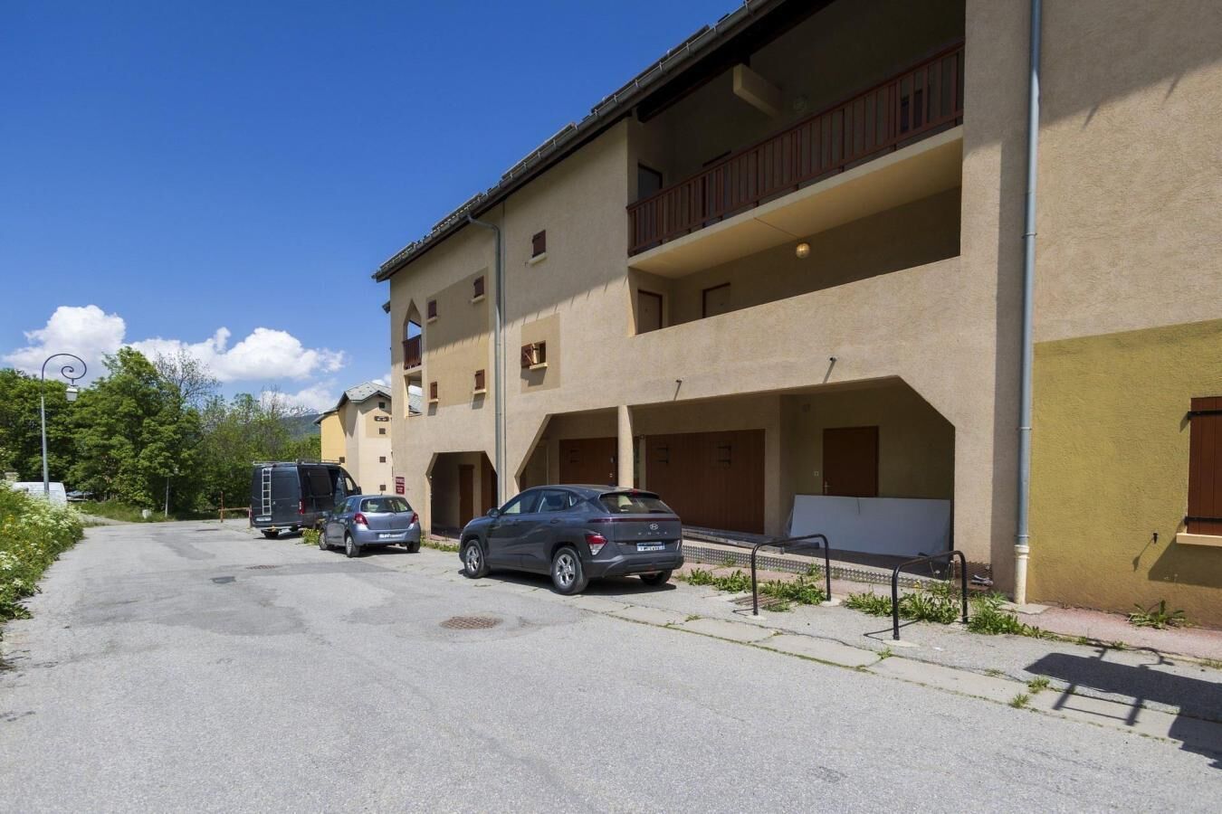 A multi-story building with balconies, a parking area with cars, and a clear sky.