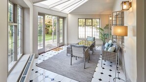 A sunlit room with large windows, a central seating area with wicker furniture, and a patterned floor.