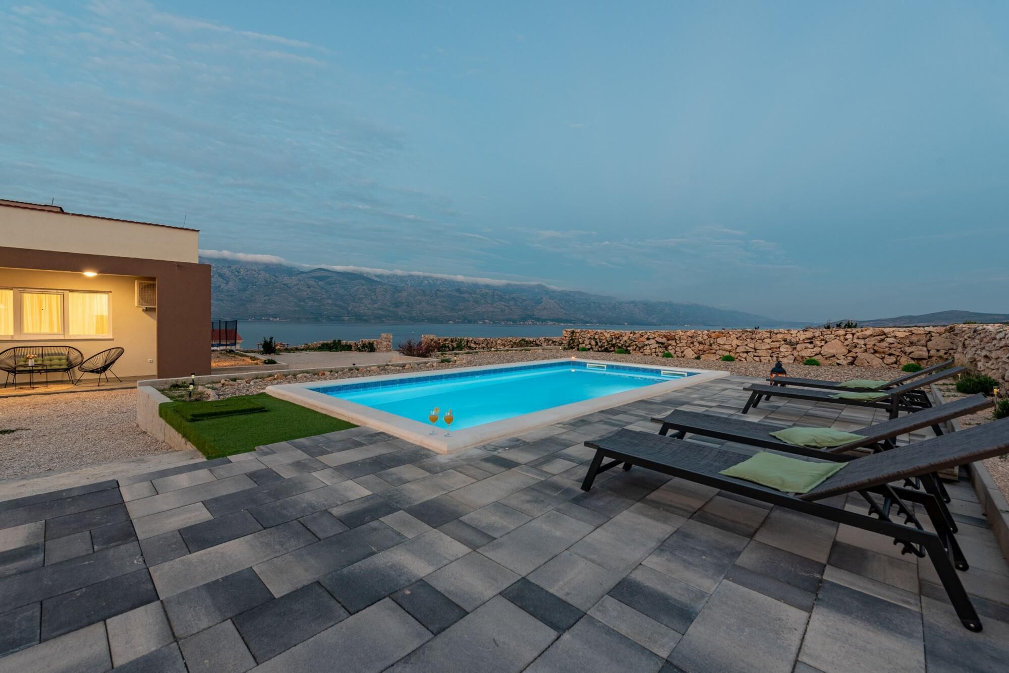 Area piscina moderna all'aperto con parete in pietra, lettini e vista sul mare.