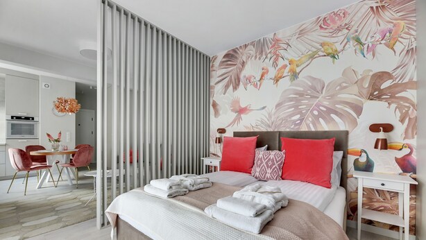 Une chambre moderne avec un grand lit, une table de chevet, un mur recouvert d’un papier peint à motifs d’oiseaux tropicaux et un coin repas avec une chaise rouge.