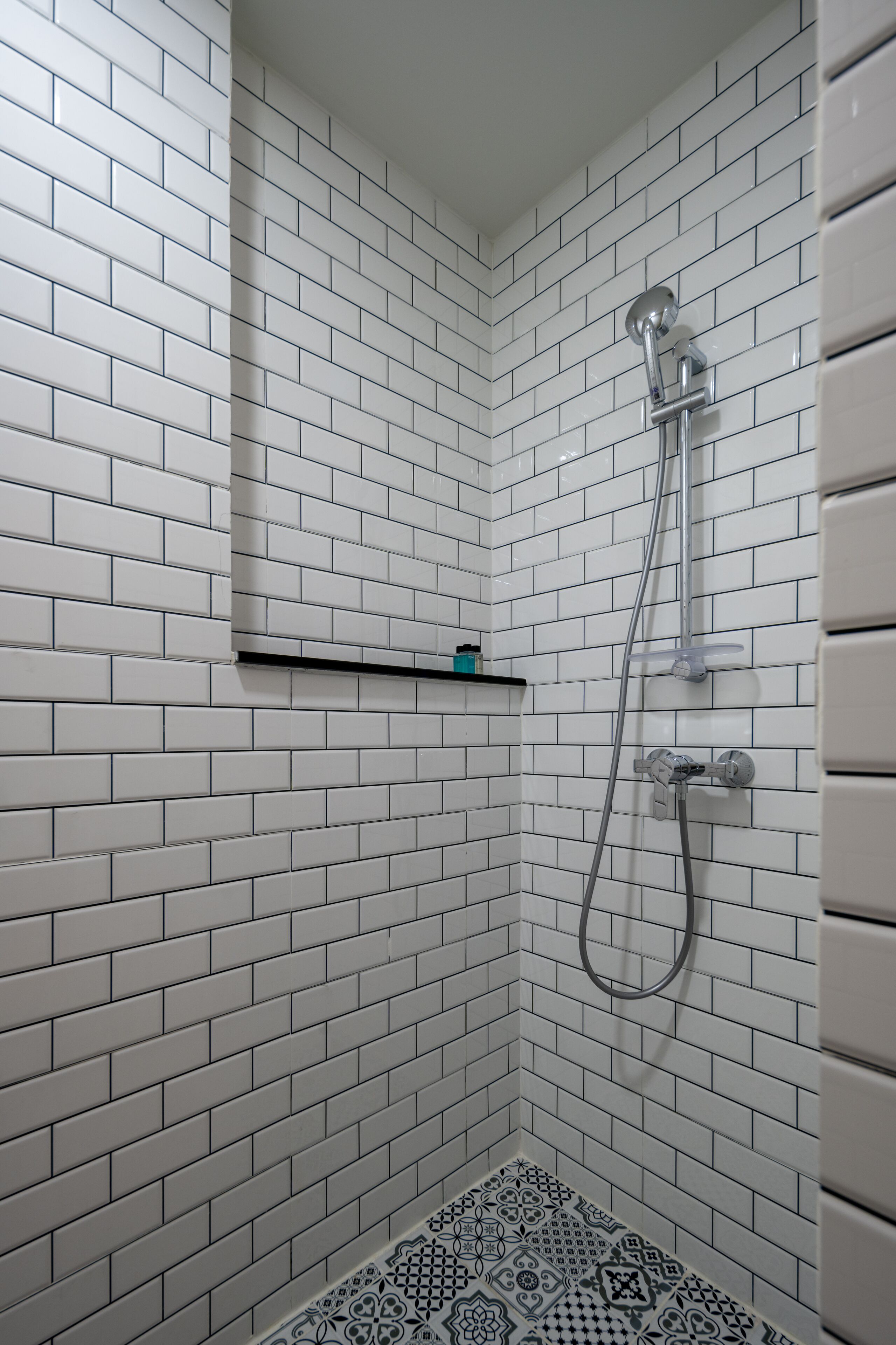 A shower area with white subway tiles on the walls and floor, a showerhead, and a small shelf with a single item.