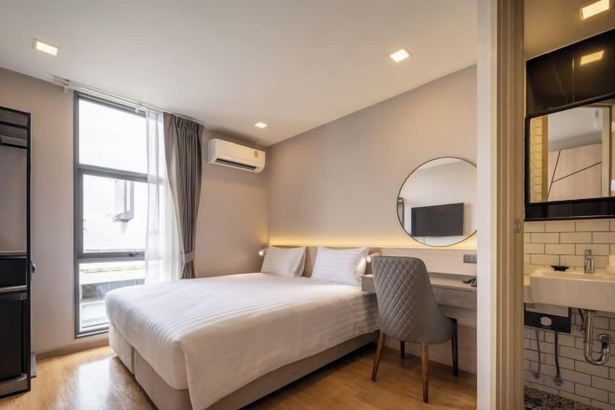 A hotel room with a bed, a desk, a chair, a window, and a bathroom with a mirror and sink.
