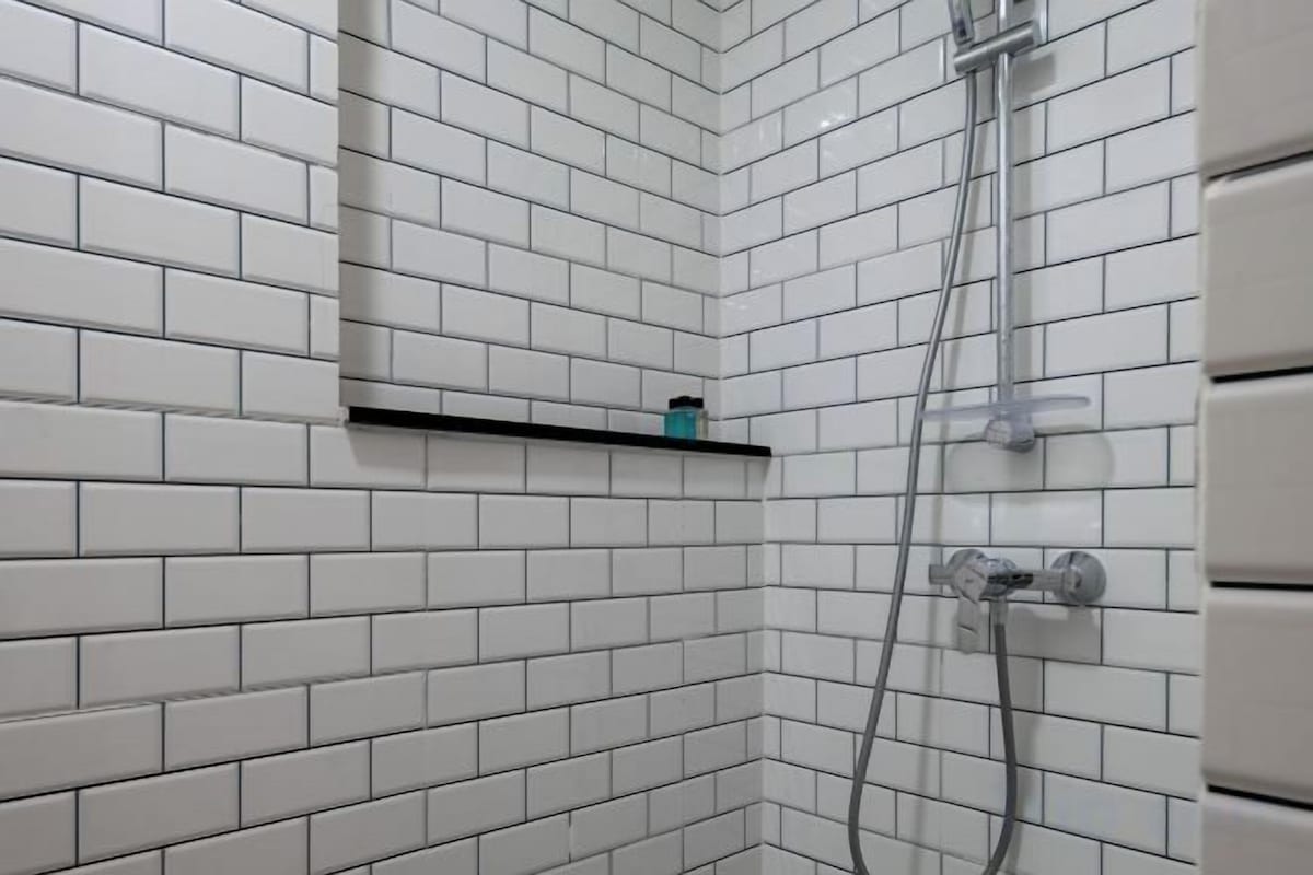 A corner of a bathroom with white tiled walls, a showerhead, and a shelf with a small item on it.