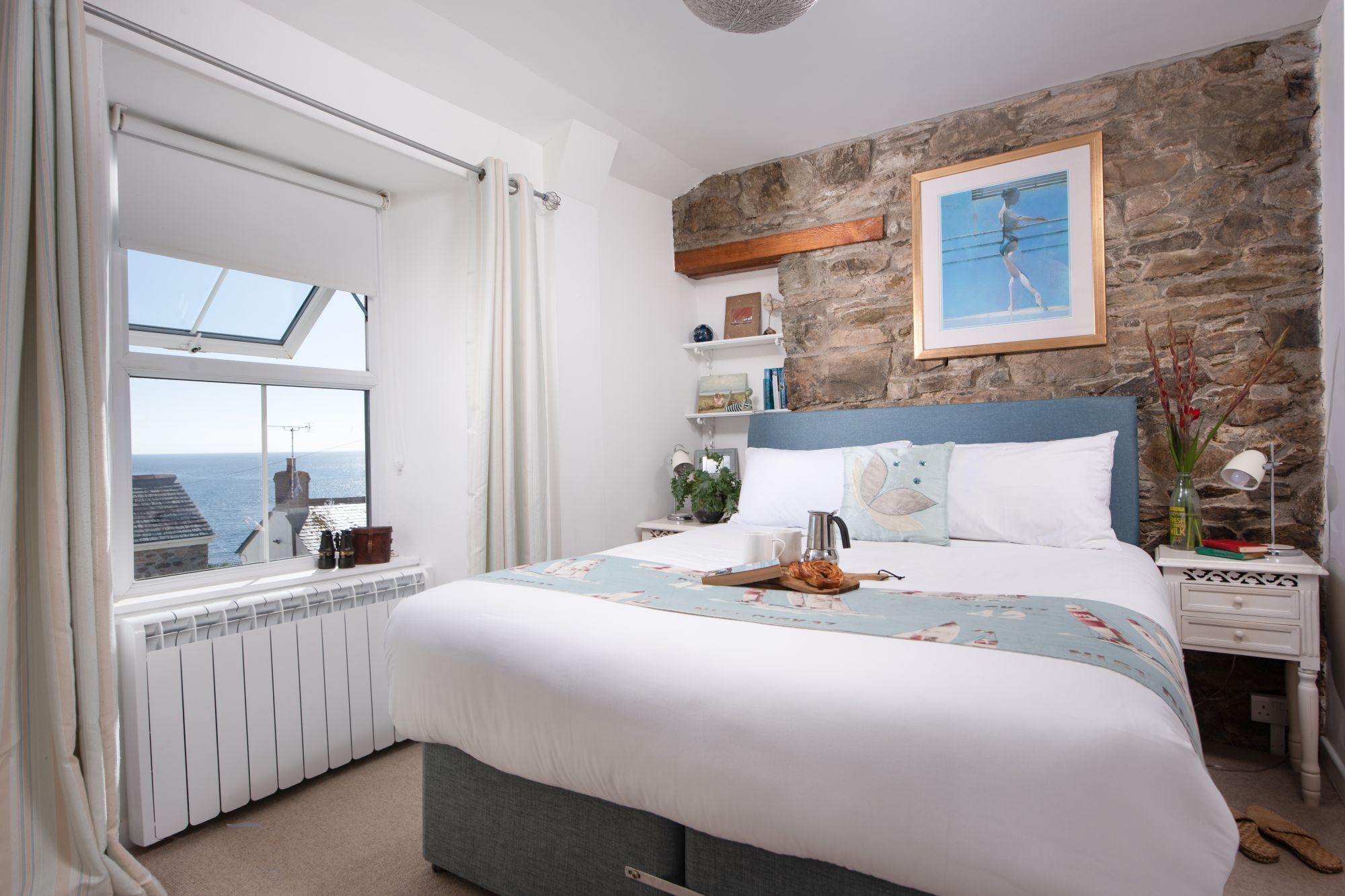 A bedroom with a large bed, a stone wall, a window with a view, and a bedside table with a lamp.