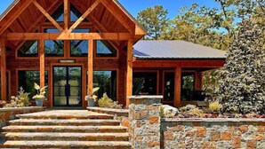 A wooden lodge with a steeply pitched roof, stone steps leading to a glass door, and a stone wall with a flower bed in front. - Grand Overlook Resort | Luxury Pool Retreat (Broken Bow)