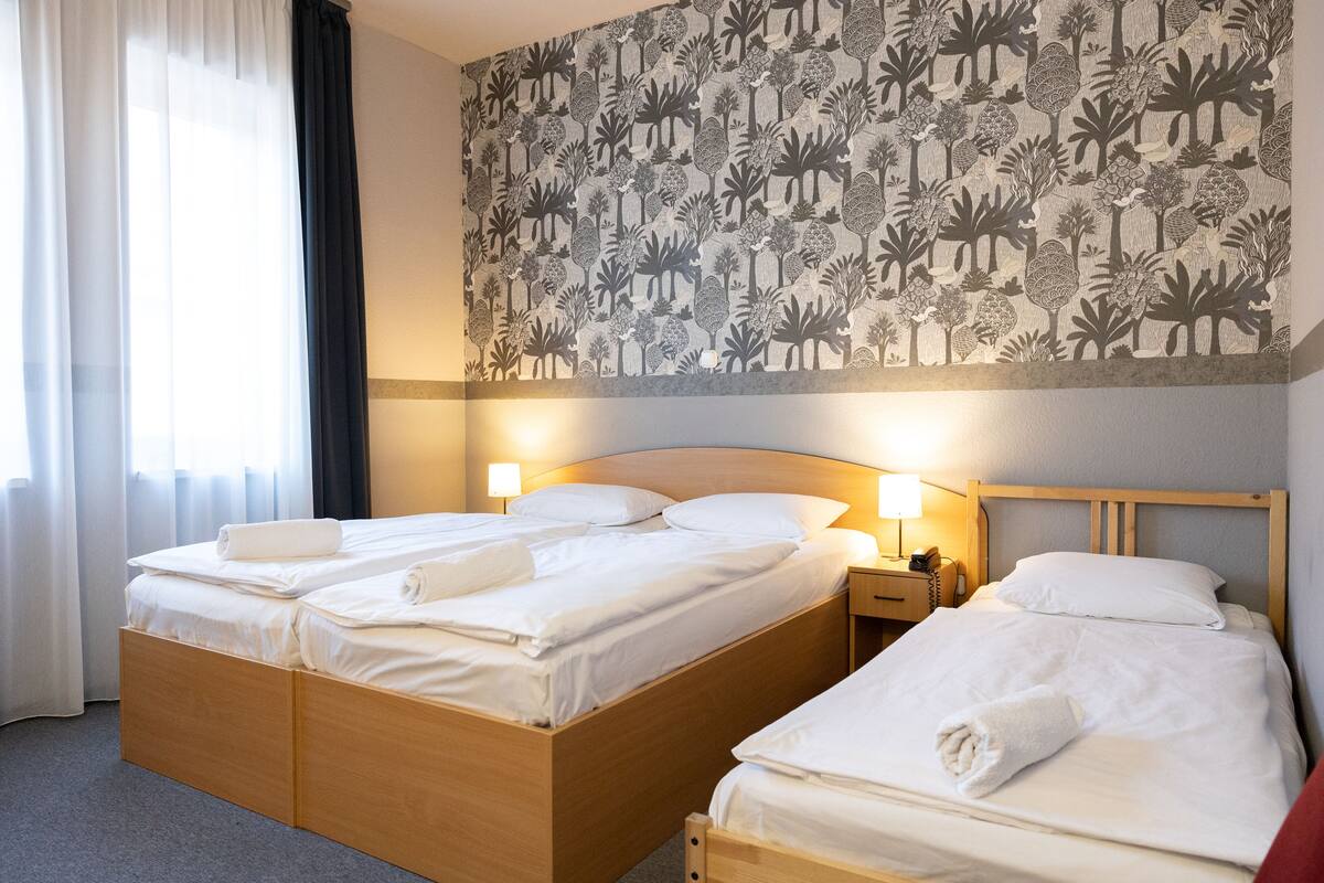 A hotel room with two beds, a wooden headboard, a nightstand with a lamp, and a floral-patterned wall.