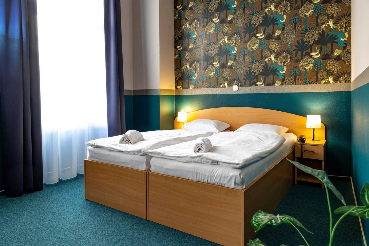 A hotel room with a wooden bed, two bedside tables with lamps, a patterned wall, and a window with curtains.