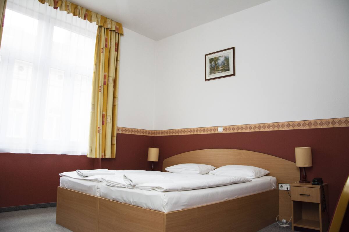 A hotel room with a wooden bed, two bedside lamps, a nightstand, a framed picture on the wall, and a window with curtains.