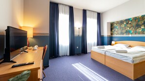 Una habitación de hotel con una cama, un escritorio con televisor, una lámpara y un cuadro en la pared.