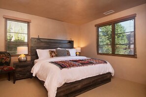 A bedroom with a wooden headboard, a bed with white bedding, two bedside lamps, a nightstand, a chair, and a window with blinds.