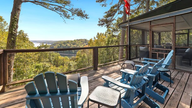 A deck with Adirondack chairs, a table, and a view of a lake and forest.