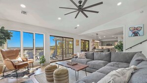 A spacious living room with a gray sectional sofa, a leather armchair, and a coffee table. There is a ceiling fan, a large window with a view, and a kitchen area in the background.