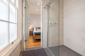 Un baño moderno con cabina de ducha de vidrio, suelo de madera, y una habitación visible con cama y mesita de noche.