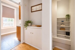 Un pasillo con una puerta blanca, una planta en maceta, un cuadro enmarcado y un estante con diversos objetos.