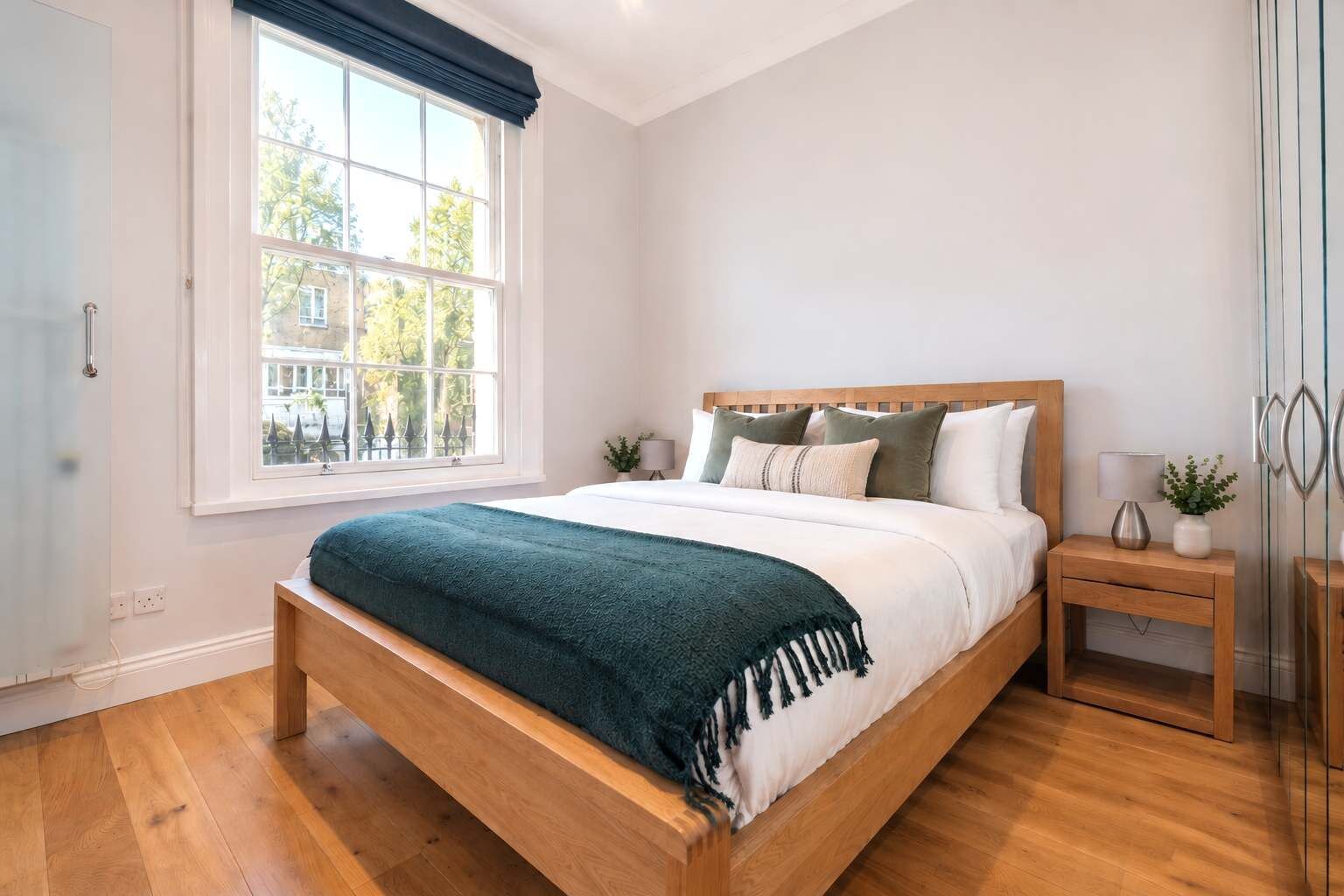 Ein Schlafzimmer mit großem Fenster, einem hölzernen Bett, einem Nachttisch mit Lampe und einem Beistelltisch mit Pflanze.