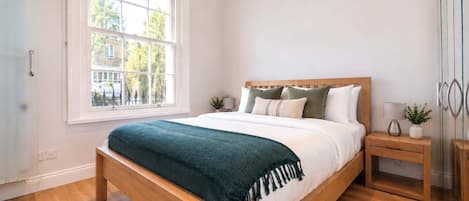 A bedroom with a large window, a wooden bed, a bedside table with a lamp, and a nightstand with a plant.