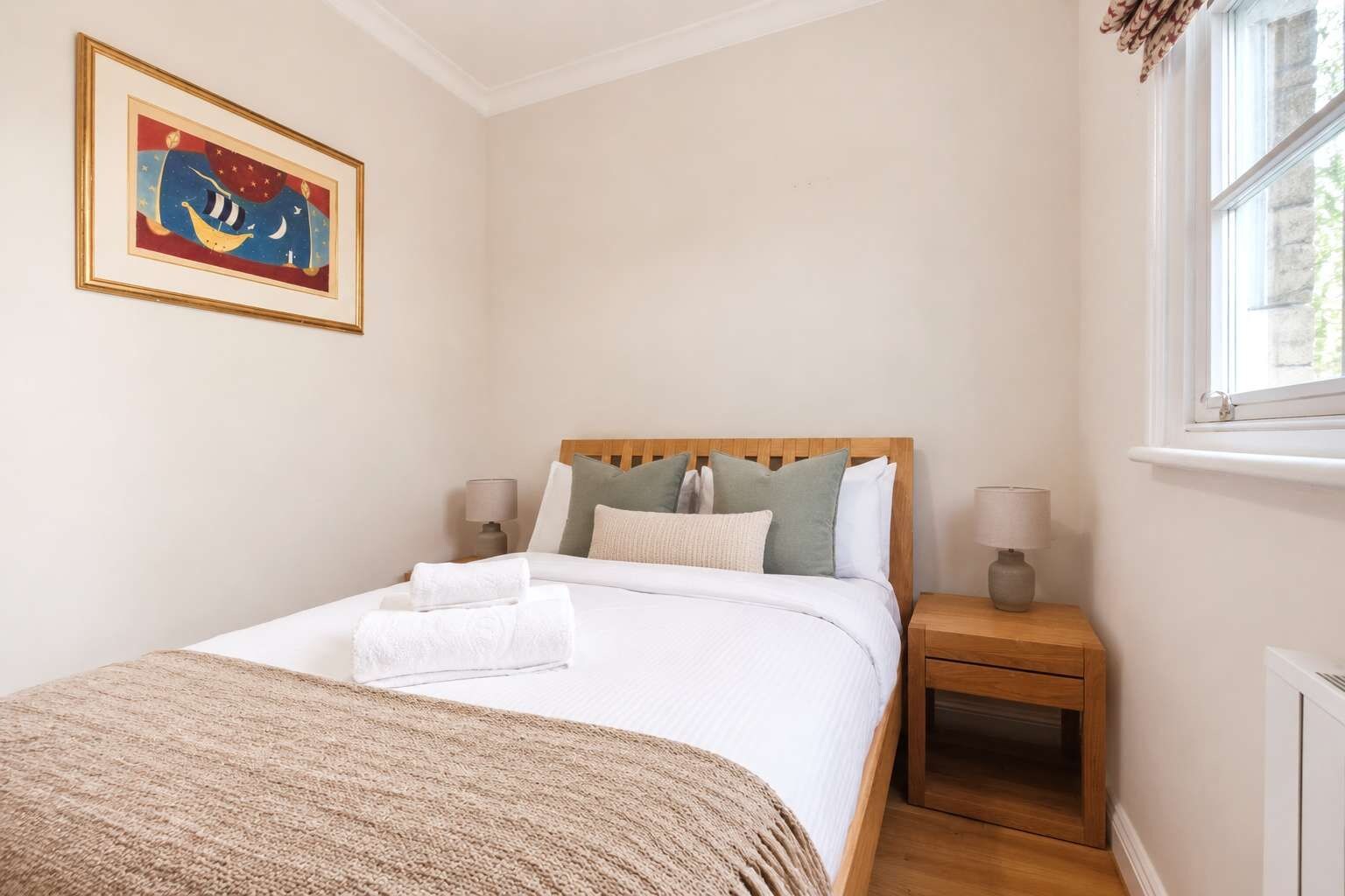 Una cama bien hecha con almohadas y una manta, una mesita de noche con una lámpara, un cuadro en la pared y una ventana con cortinas.