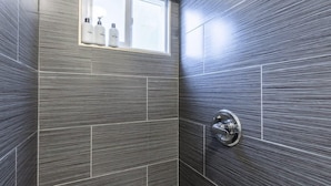 A bathroom with dark, textured tiles, a window with bottles on the sill, and a shower control panel. - @ Marbella Lane - Adorable and Quiet Studio (San Mateo)