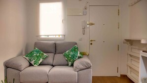 A neutral-toned sofa with two green leaf-patterned pillows, a white door with a brass handle, a white wall-mounted telephone, and a wooden floor. - Coqueto Apartamento a 5 Minutos del Retiro (Madrid)