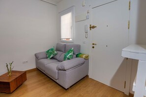 A small, well-lit room with a grey sofa, a wooden side table, and a white door with a telephone and intercom. - Coqueto Apartamento a 5 Minutos del Retiro (Madrid)