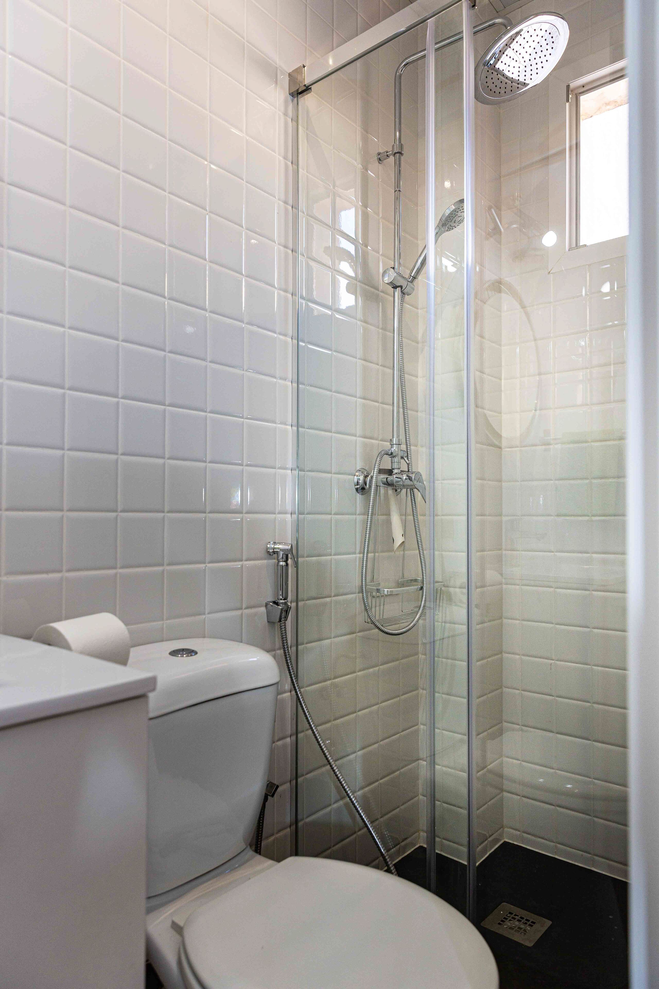 Une salle de bain avec des murs recouverts de carreaux blancs, une douche à parois de verre, des toilettes et un lavabo.