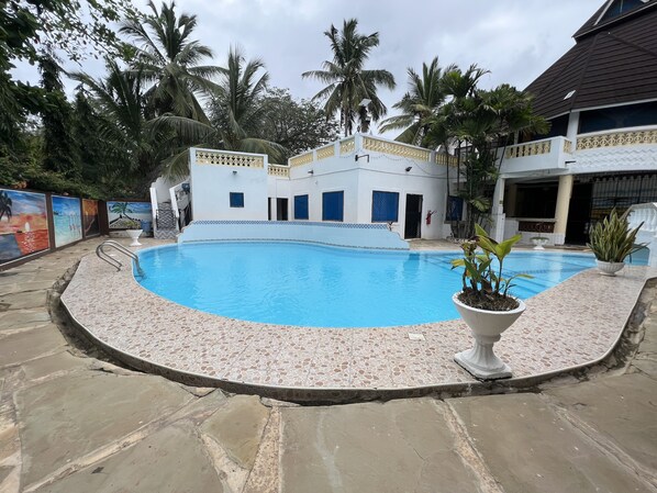 A swimming pool with a tiled edge, surrounded by a stone patio. There are palm trees and a building with a white facade and blue accents in the background.