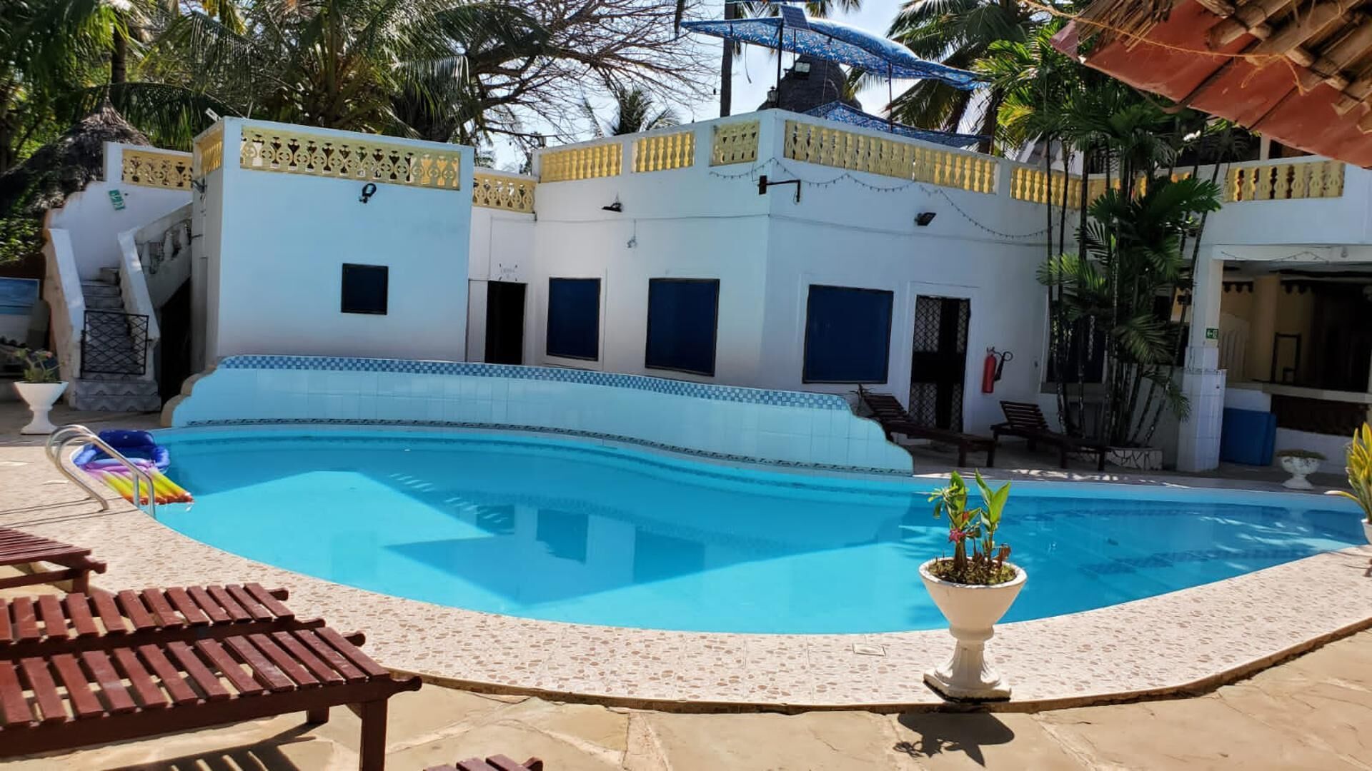 Una piscina con borde de baldosas, sillas de descanso y un edificio con varias ventanas y puertas.