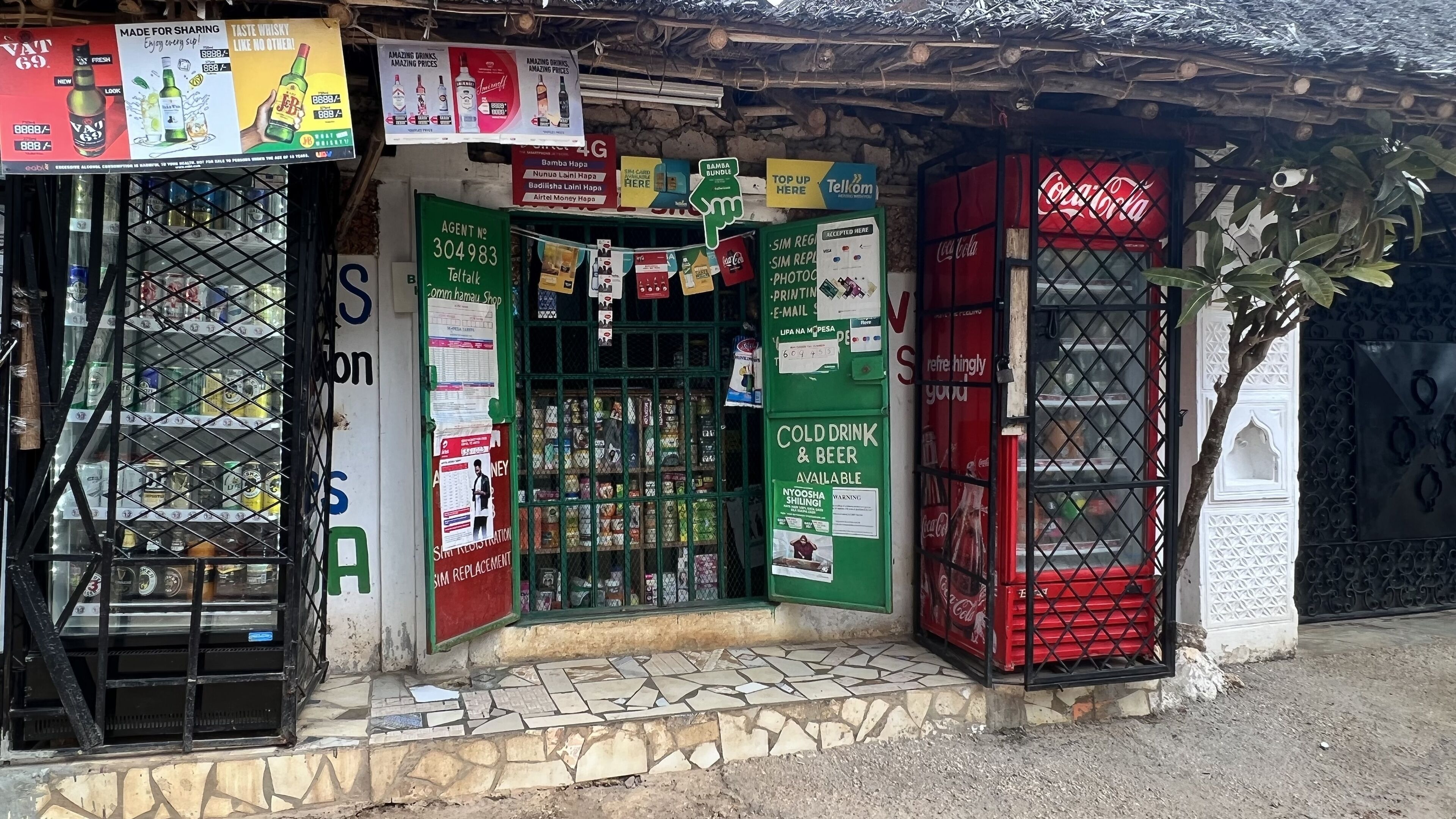 A small shop with a green door, various signs advertising drinks and services, and a Coca-Cola sign on the right.
