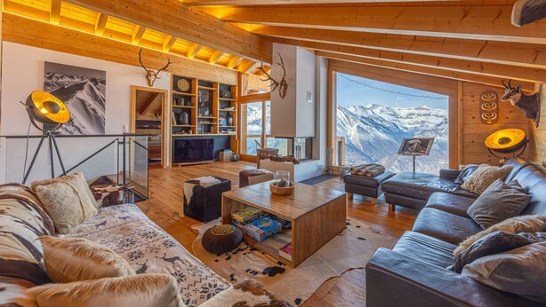 Een gezellige woonkamer met een houten plafond, een groot raam met uitzicht op de bergen, een open haard en een boekenkast.