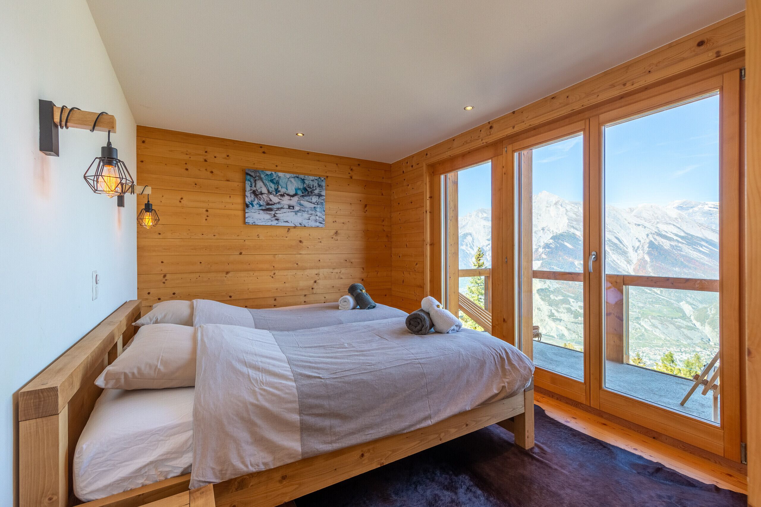 A wooden cabin room with a bed, a painting, and large windows with a mountain view.