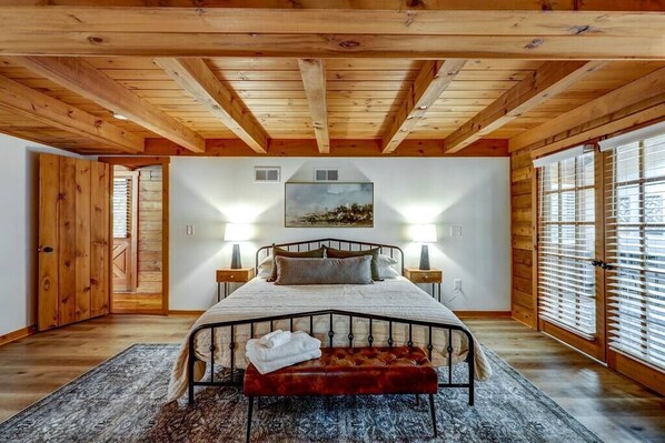 Un dormitorio con techo de madera, una cama con cabecera, dos lámparas de noche, un ventanal con persianas y un cuadro en la pared.