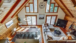 A spacious loft with a high ceiling, wooden beams, a dining area with a large table and chairs, a sofa set, a television, and a fireplace.