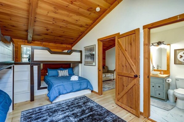 A bedroom with a bunk bed, a wooden ceiling, a bathroom with a sink and toilet, and a large wooden door.
