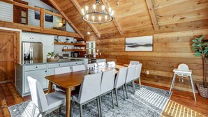 A wooden cabin interior with a dining area, kitchen, and a high ceiling with exposed beams.