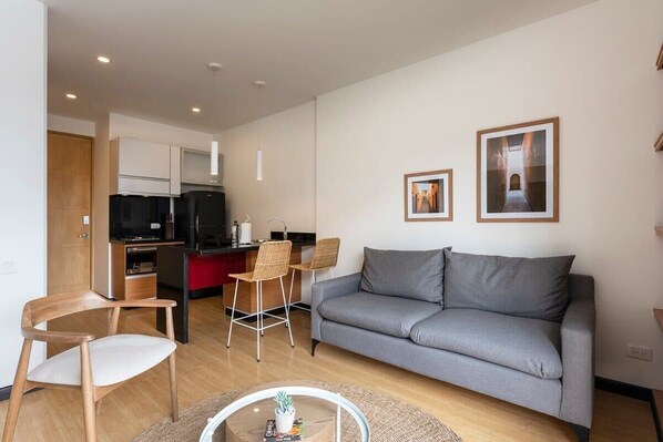 A modern living room with a grey sofa, wooden chairs, a small coffee table, and a kitchen area with a refrigerator and dining table.