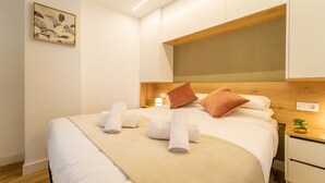 A neatly made bed with rolled towels, a wooden headboard, a bedside table with a plant, and a wall-mounted light fixture. - Estudio Prime (Alicante (Alacant))
