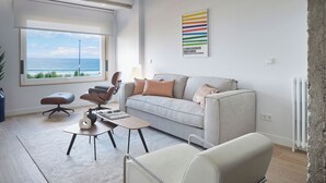 Ein modernes Wohnzimmer mit großem Fenster und Meerblick, ein graues Sofa, ein brauner Sessel und ein kleiner Tisch.