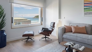 Ein modernes Wohnzimmer mit großem Fenster zum Strandblick, ein graues Sofa, ein schwarzer Bürostuhl und ein kleiner Couchtisch mit einer Zeitschrift.