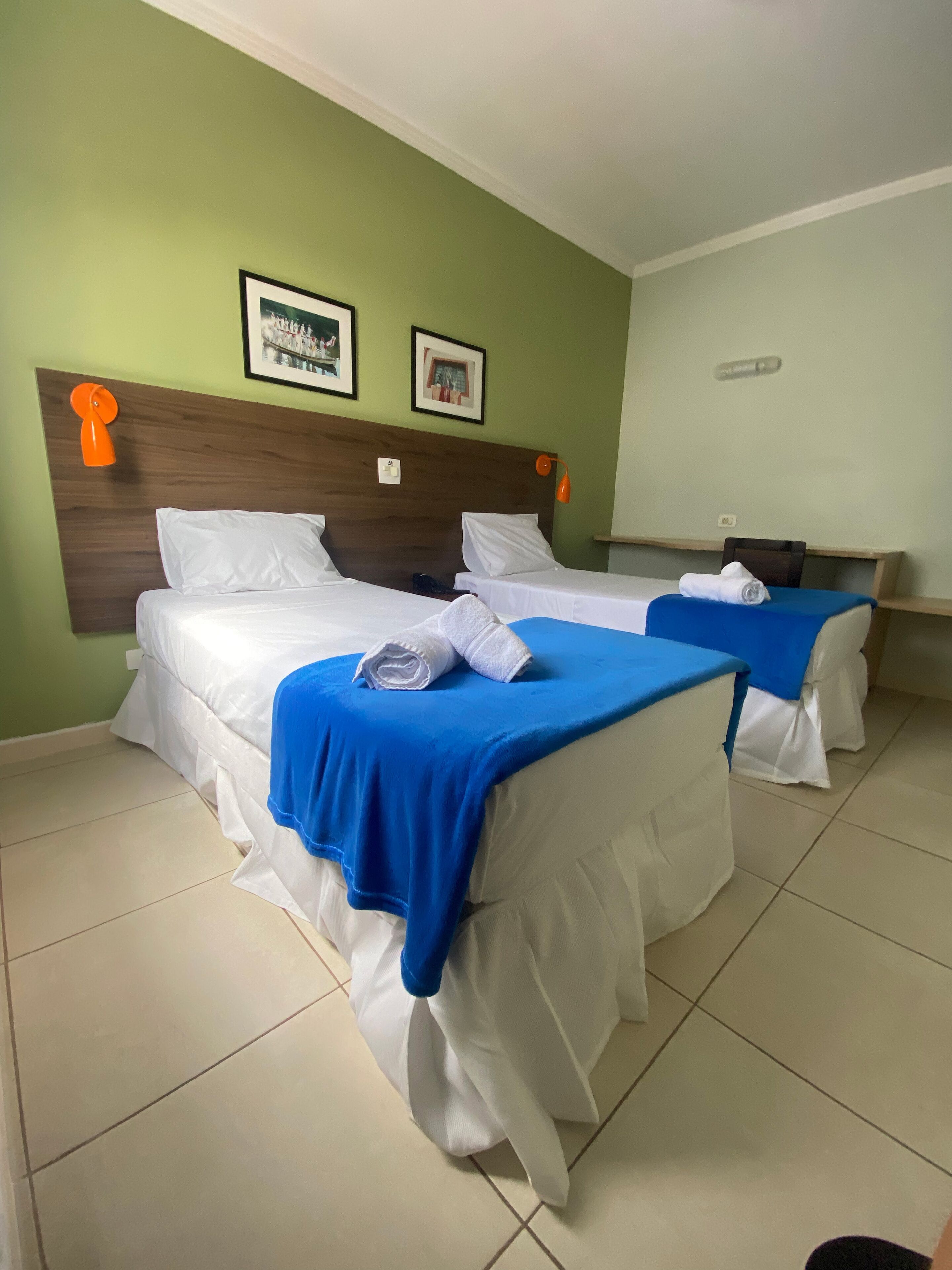 A hotel room with two beds, each with white linens and blue bedspreads. There are two framed pictures on the wall above the beds.
