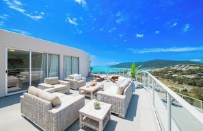 A balcony with wicker furniture, a glass door leading to an indoor area, and a view of a coastal landscape.