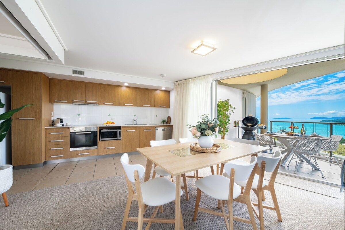 Une cuisine moderne avec des meubles en bois, un coin repas avec une table en bois et des chaises blanches, et un balcon avec vue sur la mer.
