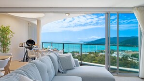 A modern living room with a large sofa, a dining area with a table and chairs, and a balcony with a view of the sea and mountains.