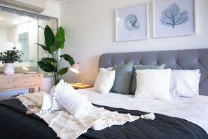 A neatly made bed with a black quilt, white pillows, and a white blanket with a lace edge. There is a bedside table with a lamp and a plant.