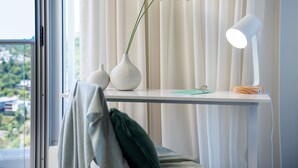 A room with a white desk, a potted plant, a lamp, and a view of greenery outside.