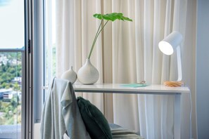 Una habitación con un escritorio blanco, una planta, una lámpara y vistas por la ventana.